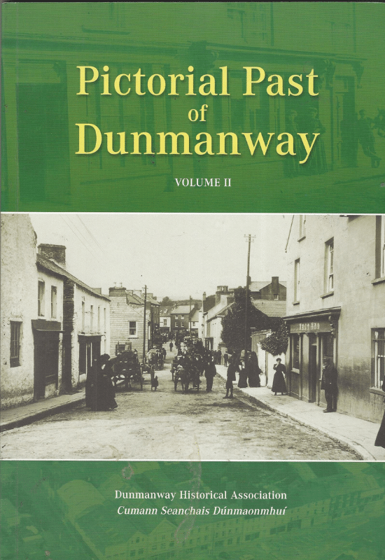 Book cover for "Pictorial Past of Dunmanway VOLUME II". The top half is dark green with gold text. Below is a large sepia photograph of an old street scene in Dunmanway with horse-drawn carts, people in period clothing, and stone buildings along a dirt road. At the bottom, in white text, are "Dunmanway Historical Association" and "Cumann Seanchais Dúnmaonmhuí" against a green background.