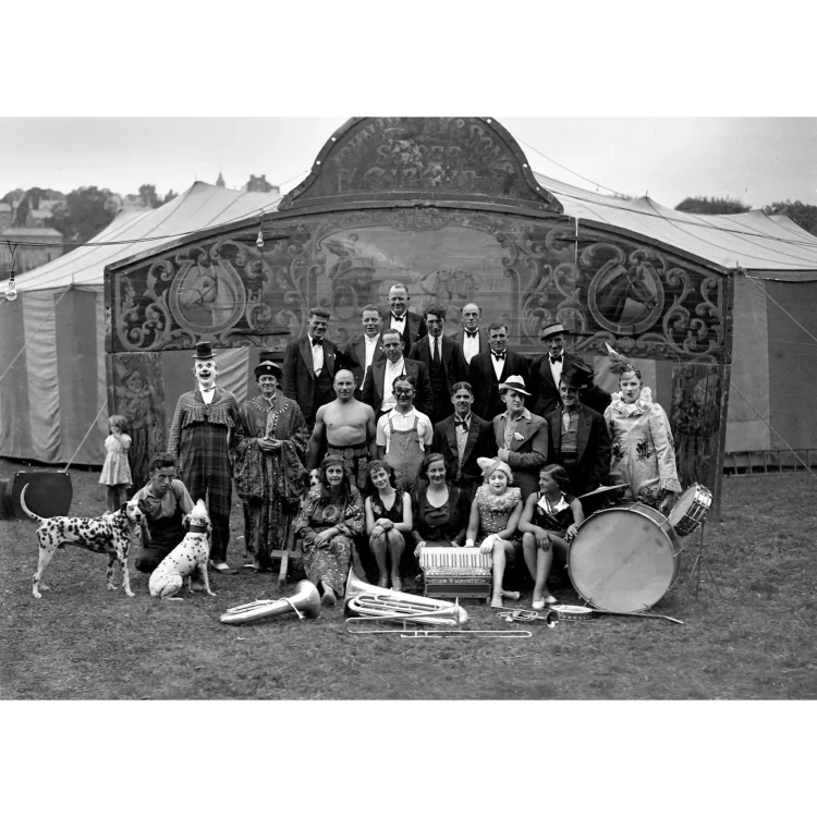 "A black and white photograph of John Duffy Circus posed together outdoors on a grassy field in front of a circus tent. The group consists of approximately twenty-four performers, including men, women, and a small child, displaying a wide variety of circus attire. In the back row, several men stand dressed in formal evening wear, including dinner jackets and bow ties. The middle and front rows feature a diverse assortment of characters: a clown in a checkered suit and top hat, a person in elaborate, heavily decorated traditional robes, a shirtless strongman displaying his muscular build, a performer in striped overalls and thick spectacles, several women in patterned dresses or performance leotards, and a white-faced clown in an ornate, ruffled costume. A small girl in a light-coloured dress stands to the far left.In the lower foreground, two dalmatian dogs are present; one stands on the left while the other sits next to a seated man. A collection of brass musical instruments, including a tuba and trombone, lies on the grass alongside a large bass drum and a snare drum on a stand.The background features an ornate, painted wooden circus façade serving as an entrance archway, decorated with illustrations of horses inside horseshoes and central carriage imagery. Behind this structure stands a large, light-coloured canvas circus tent (big top)."