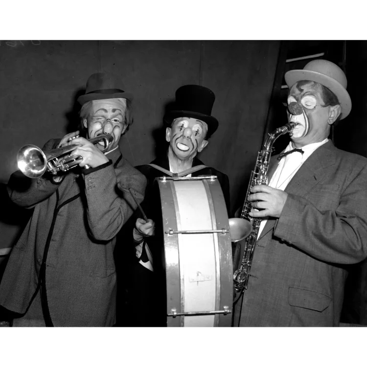 A close-up black and white photograph featuring three clowns from John Duffy’s circus performing with musical instruments against a plain, dark canvas background.The clown on the left is wearing a dark, checked jacket with contrasting trim and a fedora hat. His face is painted with exaggerated eyebrows, a large nose, and lines around his mouth; he is winking one eye while playing a silver trumpet.The central clown stands slightly behind a large marching bass drum. He wears a dark top hat and features classic sad-clown white makeup around his eyes and a wide, painted-on downturned mouth. He holds a drumstick in his hand.The clown on the right is dressed in a light-coloured, textured suit jacket with a white shirt and a dark bow tie, topped with a light bowler hat. His face paint includes white patches around his eyes and mouth, a dark red bulbous nose, and heavy dark lines down his cheeks. He is actively playing a saxophone.