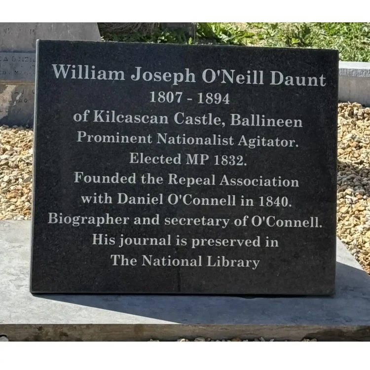 A close-up photograph of a rectangular black granite plaque placed on a grave. The crisp, white inscribed text reads: "William Joseph O'Neill Daunt, 1807 - 1894, of Kilcascan Castle, Ballineen. Prominent Nationalist Agitator. Elected MP 1832. Founded the Repeal Association with Daniel O'Connell in 1840. Biographer and secretary of O'Connell. His journal is preserved in The National Library". The plaque sits on stone gravel with green grass blurred in the background.