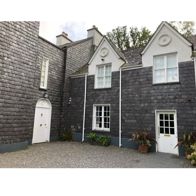 A photograph of a courtyard area at Derrynane House in County Kerry, the historic home of Daniel O'Connell. The building's exterior walls are distinctively clad in dark grey, slate-like shingles. The structure features white-trimmed sash windows, white gables with circular ornamental reliefs, and two white wooden doors leading onto a grey gravel courtyard. Potted plants flank one of the doors.