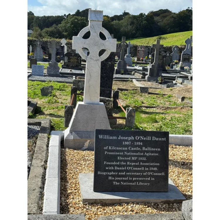 A photograph of the grave of William Joseph O'Neill Daunt in a St Patrick;s Cemetery in Dunmanway. A tall, light-coloured stone Celtic cross stands prominently as the main headstone inside a gravelled grave plot bordered by concrete. At the foot of the cross sits the rectangular black granite plaque detailing his life, accomplishments, and connection to Daniel O'Connell. Other headstones and a green sloping hill are visible in the background.