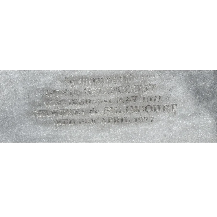 A close-up photograph of an inscription carved into a weathered, grey stone surface. The text reads: "In Memory Of GUY de SELINCOURT WHO DIED 21st MAY 1971 GEOFFREY de SELINCOURT DIED 19th APRIL 1977". The lettering is styled in a classic serif font, slightly worn by exposure to the elements.