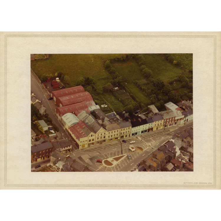 A high-angle aerial photograph displaying the reorganised Dunmanway Market Square after the Market House was demolished. The centre of the wide street has been transformed into an open parking plaza with painted white lines, featuring a new triangular concrete traffic island with a small garden bed. The square is enclosed by the multi-storey John Atkins shopfront buildings and red-roofed warehouses on one side, and a row of colourful terraced townhouses on the other.