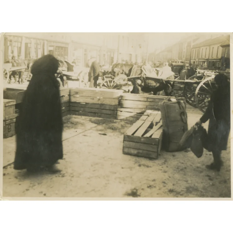A sepia-toned photograph showing a specific trading area during Market Day in Dunmanway on 3rd February 1942. A woman wrapped in a long, thick dark cloak or shawl stands on the left, looking toward a temporary livestock or goods enclosure made from low wooden slatted crates. On the right, another person carrying bundles walks past a large burlap sack resting on the ground. In the blurred background, other traders, horses, and carts line the busy street.