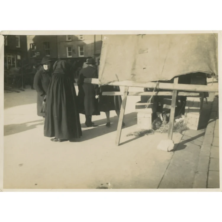A sepia-toned candid photograph from Market Day in Dunmanway on 3rd February 1942. The image highlights a woman standing in the left foreground wearing a traditional, heavy, dark Irish hooded cloak that falls to her ankles. She is conversing with another woman in a winter overcoat and hat. To the right, a simple market stall structure made from wooden planks and covered with a light-coloured canvas sheet stands over wooden crates and straw scattered on the ground.