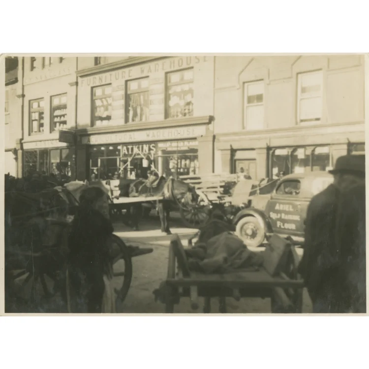 A sepia-toned street-level photograph from Market Day in Dunmanway on 3rd February 1942. In the background, the prominent signboards for "John Atkins", "China & Class", "Furniture Warehouse", and "Agricultural Warehouse" are visible above the shopfronts. A horse-drawn cart loaded with goods stands in the centre of the street, while a delivery van with "Ariel Self Raising Flour" painted on its side is parked to the right. Townsfolk move between the vehicles in the foreground.