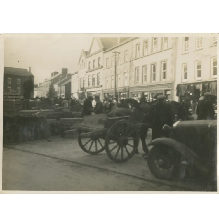 A sepia-toned historical photograph capturing a bustling scene from Market Day in Dunmanway on 3rd February 1942. In the foreground, a traditional wooden two-wheeled horse cart sits parked near the mud-splattered wing mirror of a vintage automobile. A large crowd of townsfolk, mostly men wearing caps and overcoats, fills the wide street area. They are gathered near rows of wooden crates and market goods laid out in front of a terrace of three-storey shop buildings.