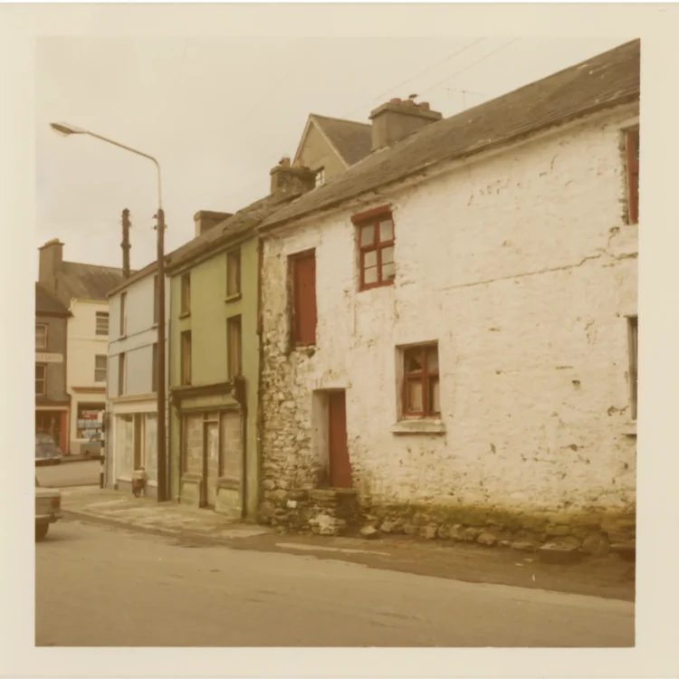 A colour photograph from the early 1970s capturing the old Market House in Dunmanway. The building's white-washed stone wall is heavily weathered and cracked, showing patches of exposed stonework near the corner. It features small timber-framed windows with red paint and a matching red wooden door on the ground floor, alongside a second-floor doorway opening directly into the air. A row of colourful terraced shops stands further down the street.