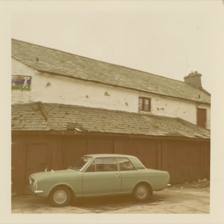 A colour photograph from the 1970s capturing a vintage light green saloon car parked in front of the old Market House building. The main building features a white-washed, heavily weathered stone exterior with a large slate roof and a single small, red-trimmed window on the upper floor. Extending forward is a lower, single-storey lean-to structure clad in dark brown vertical wooden panelling and topped with its own moss-covered slate roof. An old, faded advertisement sign is visible on the far left wall under an overcast sky.