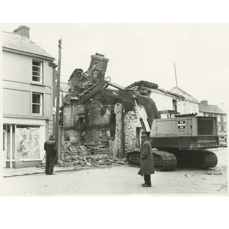 A black and white documentary photograph capturing the demolishing of Market House in 1972. A large Ruston-Bucyrus 20-H tracked excavator is positioned in the street, using its hydraulic arm to break down the stone chimney and upper roof structure of the old building. Debris and shattered masonry are piled at the base of the wall, which still shows old painted signs reading "WINE" and "SPIRITS". Two local men stand at a safe distance watching the work.