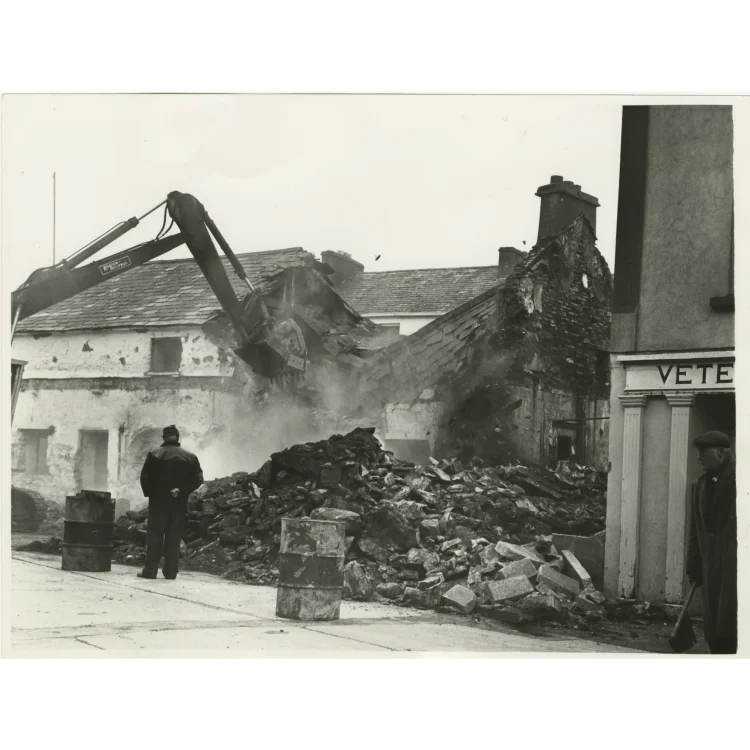 A black and white photograph documenting another angle of the demolishing of Market House in 1972. A massive pile of broken stones, mortar, and slate fills the foreground, flanked by two metal oil drums. In the background, the metal bucket of a mechanical excavator crashes through the sloped roof and stone walls of the white-washed structure, sending up a cloud of dust. A bystander with his hands behind his back watches from the left.