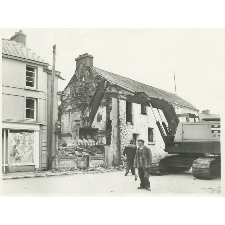 A black and white photograph capturing a later stage of the demolishing of Market House in 1972. The large Ruston-Bucyrus excavator uses its heavy bucket to tear down the front facade of the building, exposing the internal stone walls and plaster of the upper storey. Rubble from the crumbling structure spills onto the road behind the remnants of the old pub fronting that reads "WINE" and "SPIRITS". A workman stands in the foreground observing the progress.