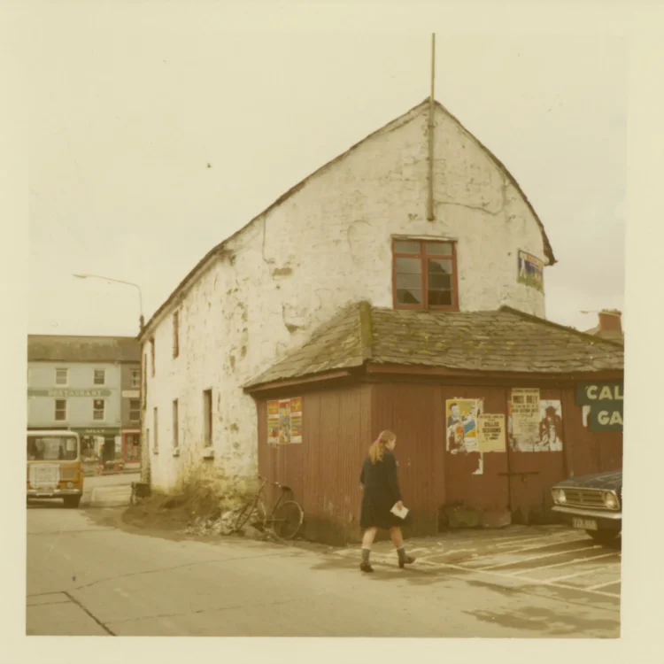 A colour photograph from the early 1970s showing the historic Market House building in Dunmanway prior to its demolition. The two-storey building has a white, rough-textured stucco exterior with a tall gabled profile and a central upper window. A lower single-storey section clad in dark reddish-brown corrugated iron extends to the right, decorated with various event posters and a "Calor Gas" sign. A young girl is walking past the building on the tarmac.
