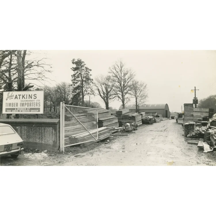 A black and white photograph showing the outdoor yard of John Atkins Timber Importers. On the left, a large white sign reads "John Atkins (Dunmanway) Ltd. Timber Importers, A Member of the James McMahon Group". An open white mesh gate leads into a wide, unpaved yard filled with large stacks of processed timber planks. A large curved Nissan-style tin shed stands in the background under a cloudy sky.