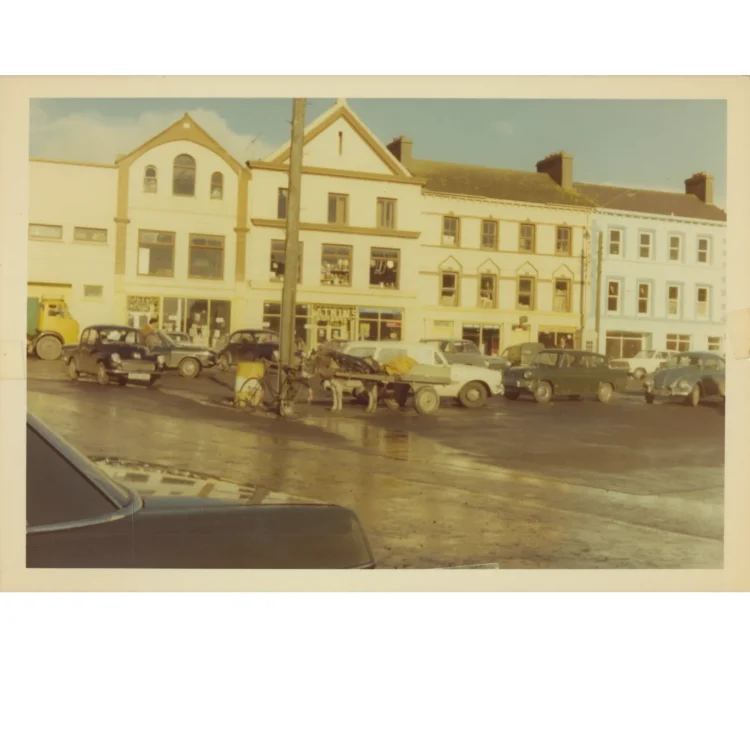 A faded colour photograph capturing a busy market day on the street outside John Atkins in Dunmanway. In the centre, a donkey harnessed to a wooden flat-bed cart stands next to a bicycle leaning against a telegraph pole. Vintage cars from the mid-20th century, including a Volkswagen Beetle, are parked across the wide street area in front of the cream and blue terraced shopfronts.