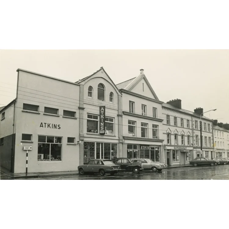 A black and white photograph depicting the full street-facing exterior of the John Atkins premises. The large, interconnected storefront spans multiple architectural styles, from a modern flat-roofed section on the left displaying the name "ATKINS", to a traditional gabled building in the centre with an "ATKINS" sign hanging vertically, and a "Supermarket" entrance on the right. Several 1970s saloon cars are parked outside on the wet tarmac.