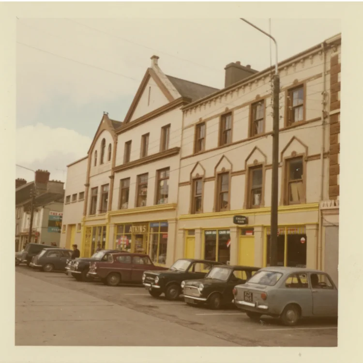 A colour photograph from the 1970s showing a street view of the John Atkins shopfront in Dunmanway. The three-storey building features a cream facade with distinct yellow trim along the windows and ground floor. Several vintage cars, including a Morris Minor and a Mini, are parked diagonally along the kerb in front of the shop. A prominent vertical sign on the shopfront reads "ATKINS".