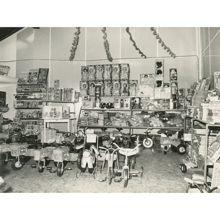 A black and white archival photograph showcasing a large Christmas toy display inside the John Atkins shop. The shelves are packed with a wide variety of boxed dolls, teddy bears, board games, and playsets. Tinsel garlands hang from the ceiling. On the floor in the foreground, a fleet of children's ride-on toys is arranged, including toy tractors, tricycles, and a ride-on donkey.