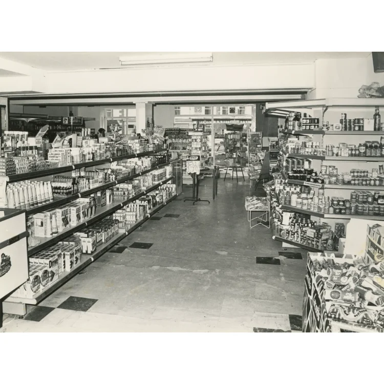 A black and white interior photograph of the modernised grocery and hardware aisles inside John Atkins supermarket. Long, well-stocked metal shelving units run down the left side, filled with household cleaning products like Ariel and Drive. The right-hand shelves are stacked with canned goods, jars, and provisions. A checkout counter or display bin filled with sweets stands in the immediate bottom right corner.