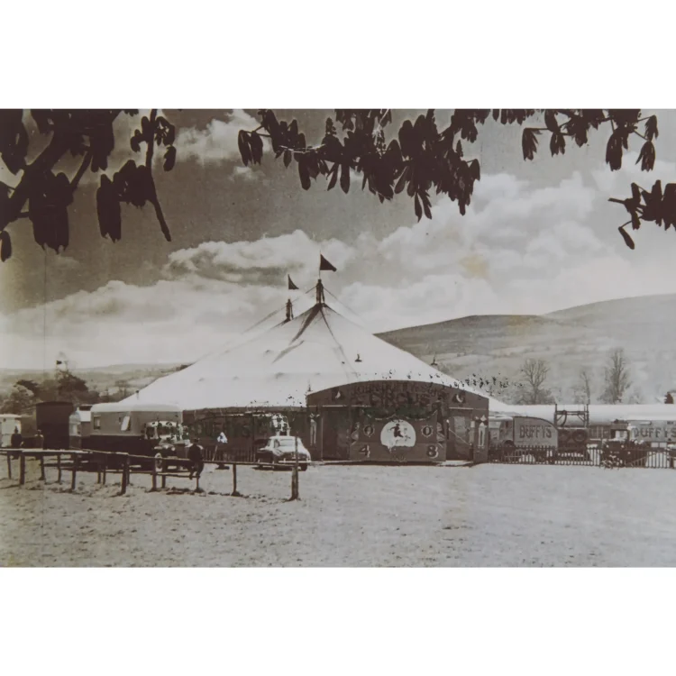 A black and white photograph of the John Duffy Circus pitched at the Showgrounds in Clonmel in 1956. The sprawling white canvas big top tent stands in the centre of an open field, adorned with small dark flags at its peaks. Several vintage living trailers, trucks, and support vehicles are parked in a neat row behind a low wooden post-and-rail fence in the foreground, with rolling hills rising in the distance.