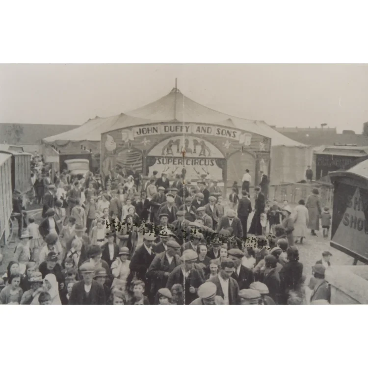 A black and white historical photograph depicting a large crowd of people gathered outside the Duffy Circus tent in Sligo in 1937. A diverse mix of adults and children dressed in early 20th-century attire fill the fairground space. In the background stands the large circus big top tent with an ornate entrance façade reading "JOHN DUFFY AND SONS SUPER CIRCUS", decorated with illustrations of performing animals and circus clowns.
