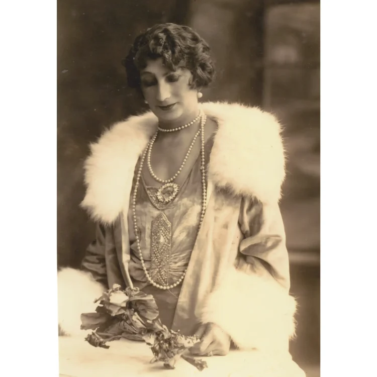 A sepia-toned vintage portrait photograph of Rosie, the fortune teller from Duffy's Circus. She is elegantly dressed in a 1920s style silk or satin dress with an intricate geometric pattern panel down the front. She wears a lavish coat with a large, fluffy white fur collar and matching cuffs. Multiple strands of long pearl necklaces drape over her chest, pinned with a prominent circular jewel brooch, and she carries a small bouquet of flowers.