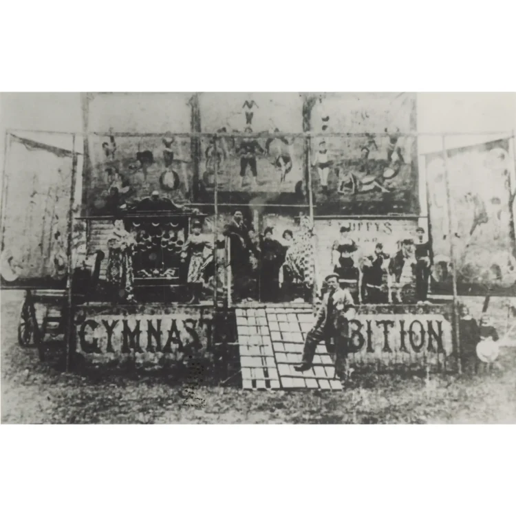 A historical black and white photograph capturing the early years of John Duffy's Circus. A troupe of performers, including clowns, acrobats in stage costumes, and musicians, are posed in a row on a elevated wooden circus wagon stage labeled "GYMNASTIC EXHIBITION". On the far left, John Duffy I stands prominently at the top of a wooden slatted ramp leading up to the stage.