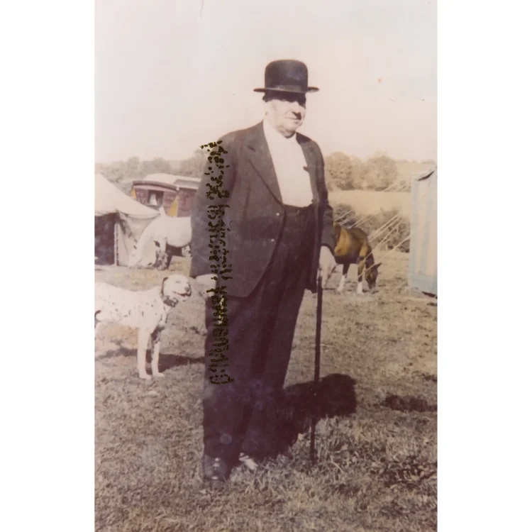 A vintage photograph of John Duffy II standing outdoors in a grassy field. He is dressed formally in a dark three-piece suit, a crisp white shirt, and a black bowler hat, while holding a dark walking cane in his left hand. In the background, circus horses graze freely near living wagons and canvas tents, capturing the atmosphere of the traveling show.