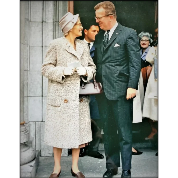 A colour photograph capturing a candid moment between American political strategist Larry O'Brien and his maternal aunt, Julia Sweeney, standing outdoors next to a stone pillar at Cork City Hall during a visit in 1963.On the left, Julia Sweeney stands facing right, smiling warmly at her nephew. She wears a structured, light beige or cream-coloured tweed coat that falls below the knee, featuring prominent brown buttons. Her outfit is completed by a matching folded fabric hat, white gloves, and brown, low-heeled shoes. She holds a small white paper or card in both hands and has a dark brown leather handbag tucked under her left arm.On the right, Larry O'Brien stands facing left, looking down at his aunt with a smile. He wears a dark, pinstriped navy or charcoal suit with a white shirt, a dark patterned tie with white polka dots, a white pocket square, and dark dress shoes. He also wears his characteristic thick-rimmed glasses.In the background, a man in a dark suit and a woman with grey hair wearing glasses are partially visible, looking toward the pair. The architectural setting consists of the large, light-grey stone blocks and a classic column base of Cork City Hall on the far left.
