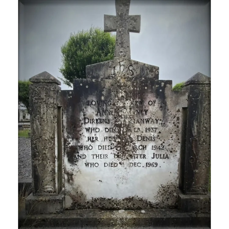 A grey, weathered stone headstone of Larry O’ Brien’s grandparents and Aunt, Julia Sweeney’s grave positioned in an outdoor churchyard against a cloudy, overcast sky. The headstone features a symmetrical design with a central rectangular plaque topped by a stone cross. The cross has the stylised letters "IHS" carved into its base block. The central plaque is flanked by two decorative side pillars with geometric caps and carved floral or leaf motifs running vertically down their fronts. The stone shows significant dark weathering, lichen, and staining across its surface, particularly on the top and left sides. Green foliage and trees are visible in the soft-focus background.
