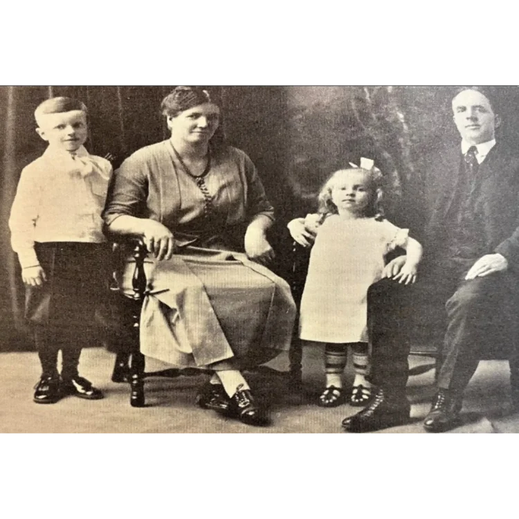 A black and white studio family portrait of Larry O’Brien posed against a dark, neutral backdrop. On the far left, Larry O’ Brien stands as a young boy stands facing forward, wearing a light-coloured, long-sleeved shirt with a wide collar and dark shorts or trousers that reach the knee, paired with dark socks and shoes. Seated next to him in a dark wooden chair is his mother facing forward with a calm expression. She wears a dark, loose-fitting dress with a long necklace.To her right stands his sister wearing a light-coloured, short-sleeved dress with dark, striped socks and dark shoes. She has a bow in her hair.On the far right, Larry O’Brien’s father sits facing slightly inward toward the family. He wears a dark three-piece suit consisting of a jacket, waistcoat, and trousers, with a stiff white shirt, a dark tie, and dark lace-up boots.