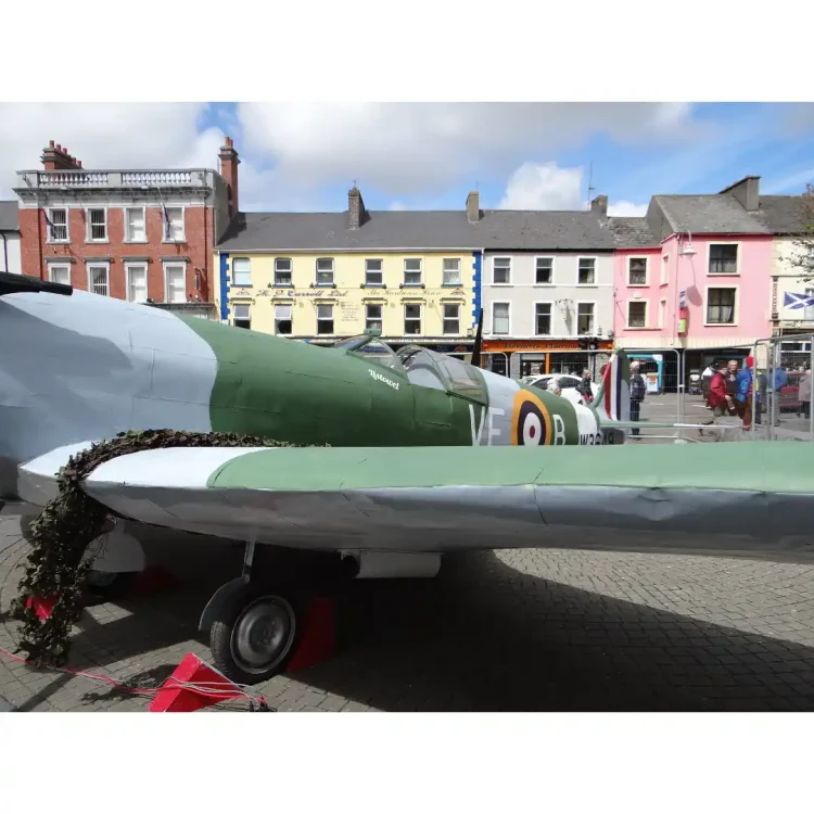 A close-up of a full-scale replica or restored historical fighter aircraft, painted in a military green and grey camouflage scheme. The aircraft is parked outdoors on a grey herringbone-patterned brick pavement in the Square in Dunmanway.The side of the fuselage prominently features the large, white letters VF to the left of a classic Royal Air Force (RAF) roundel target symbol (consisting of concentric rings of yellow, blue, white, and a red center). To the right of the roundel is a large, white letter B, followed further back towards the tail by the serial number W3649 in black text.The cockpit canopy is visible, showing a clear windscreen structure. On the front section near the cockpit, the word Listowel is written in a white script font. A section of dark green camouflage netting is draped over the wing root on the left side.In the background, across the square, is a row of multi-storey town buildings . The buildings include a red-brick structure with white window frames and an inscription for "AIB Bank", and a bright yellow building next to it featuring commercial signage. A white temporary barrier stands on the pavement behind the aircraft.