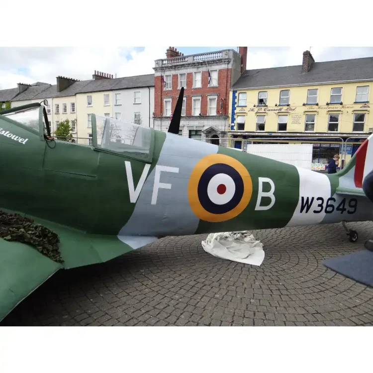 A close-up, side-angle view of a full-scale replica or restored historical fighter aircraft, painted in a military green and grey camouflage scheme. The aircraft is parked outdoors on a grey herringbone-patterned brick pavement in the Square in Dunmanway.The side of the fuselage prominently features the large, white letters VF to the left of a classic Royal Air Force (RAF) roundel target symbol (consisting of concentric rings of yellow, blue, white, and a red center). To the right of the roundel is a large, white letter B, followed further back towards the tail by the serial number W3649 in black text.The cockpit canopy is visible, showing a clear windscreen structure. On the front section near the cockpit, the word Listowel is written in a white script font. A section of dark green camouflage netting is draped over the wing root on the left side.In the background, across the square, is a row of multi-storey town buildings . The buildings include a red-brick structure with white window frames and an inscription for "AIB Bank", and a bright yellow building next to it featuring commercial signage. A white temporary barrier stands on the pavement behind the aircraft.
