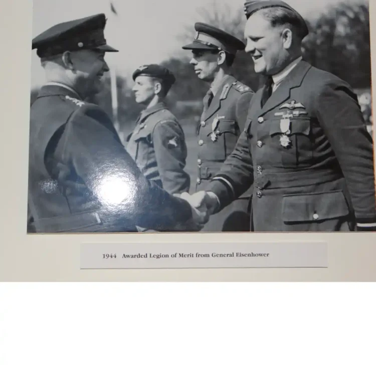 A black-and-white photograph mounted on a cream-coloured card board. The photograph depicts an outdoor military decoration ceremony in 1944.On the left, General Dwight D. Eisenhower is seen in profile from the waist up, wearing a service cap and dark winter uniform coat with stars visible on his shoulder strap. He is smiling and shaking hands with an RAF officer on the right, identified by the caption as George Beamish.George Beamish is wearing an RAF service dress uniform with pilot wings and medals pinned to his left chest pocket, smiling broadly back at Eisenhower. In the background between them, two other military personnel stand in a row at attention. Below the print, a clean white label strip contains printed black text detailing the event.