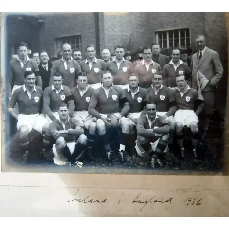 A black-and-white, vintage team photograph of an Irish international rugby squad from 1936, mounted on a historical display board. The image features nineteen men posed in three rows against the brick wall of an old stadium or pavilion building. Charles Beamish is the 3rd from right, second row seated. The players wear dark jerseys with a shamrock crest on the left breast, light-coloured shorts, and dark socks. Several individuals in the back row and on the far right stand wearing formal suits or blazers. The man sitting in the center of the middle row holds a vintage leather rugby ball.At the bottom of the mount, there is cursive handwriting in black ink identifying the match.