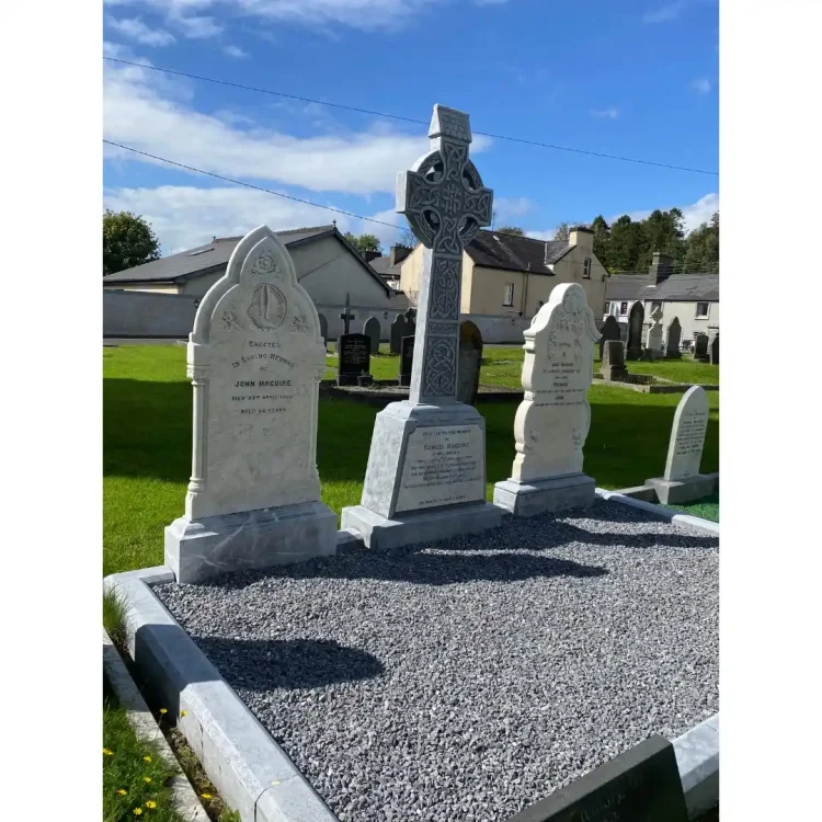 A wide, eye-level outdoor shot depicts the Maguire family burial plot enclosed by a low, grey stone border filled with fine grey gravel. The plot contains three prominent white marble and grey stone headstones situated in a green graveyard under a bright blue sky with scattered white clouds. Left Headstone: A tall, white marble monument with a decorative arched top featuring a relief carving of an anchor inside a circle. The main body of the stone is rectangular and bears an engraved inscription. Centre Headstone: A tall Celtic cross made of grey stone, adorned with intricate, traditional interlaced knotwork carvings along its shaft and face. It stands on a multi-tiered, tapered base. The front face of the base features a smooth white marble plaque with an engraved inscription. Right Headstone: A white marble headstone with a decorative, curved top mirroring the style of the left monument, featuring engraved text across its front face.In the background, there is a vibrant green lawn with several other scattered historic headstones, bounded by a low cream-coloured wall and residential buildings with slate roofs under a bright sky.