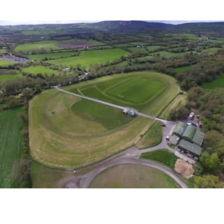 A high-angle, wide aerial drone photograph captures a green sports and racing complex surrounded by rural farmland. The central feature is a large, oval-shaped grass racetrack showing lighter green and straw-coloured texturing along the path. Enclosed within the oval track is a well-maintained, bright green rectangular football pitch with white goalposts visible at each end. A narrow grey asphalt access road bisects the oval field diagonally from the upper left to the lower right. In the lower right corner, a cluster of green-roofed agricultural or club utility buildings stands next to a small parking area with a few vehicles. The surrounding landscape consists of rolling green fields, dense hedgerows, and distant low hills under a soft, overcast sky.