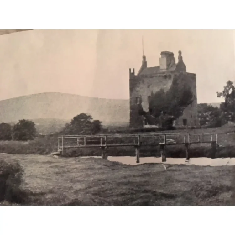 A vintage, grain-textured black-and-white photograph depicts a tall, historic stone castle tower house standing in a rural field.The rectangular castle features small windows, defensive battlements at the roofline, and a tall flagpole mounted on its top corner. A heavy growth of dark ivy covers a significant portion of its front-facing wall. In the foreground, a rustic wooden footbridge with simple railings spans across a narrow, calm river that reflects the sky. The surrounding terrain consists of grassy banks, scattered trees to the left and right, and a soft, low mountain range rising gently in the distant background under a pale sky.