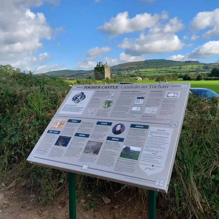 A wide shot shows a large, rectangular metal information display sign mounted on green posts in front of Togher Castle. The sign's surface is covered with comprehensive printed text columns, historical illustrations, timelines, maps, and photographic panels detailing the history of Togher Castle. The actual stone ruin of Togher Castle is visible in the distant background, centered right above the top edge of the sign, nestled amongst rolling green hills and fields under a blue sky filled with white clouds. On the left, the sign is bordered by thick green bushes and wild brambles, while a dirt path runs along the bottom left corner.