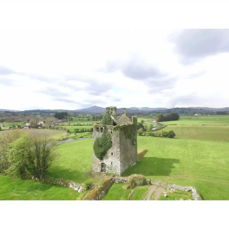 An aerial drone photograph shows Togher Castle standing in a wide, sunlit green field. The square stone tower house is positioned centrally, with thick curtains of green ivy cascading down its left and upper walls. The roof structure is open and partially ruined, exposing the interior stone dividers. A clear shadow of the tower stretches across the bright green grass to the right. In the foreground, a stone wall with a small metal gate opens onto a dirt track that leads toward the castle. The background reveals a vast, rolling landscape of hedgerows, cultivated green fields, a winding river, a small cluster of farm buildings on the left, and distant blue-grey mountains under a bright sky with scattered white clouds.
