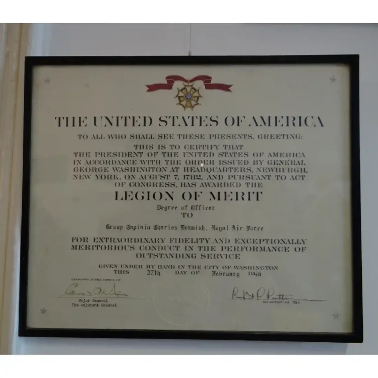 A front-on, close-up photograph shows a framed, rectangular military award certificate (Legion of Merit) presented on slightly aged, off-white paper. The document is mounted inside a simple, thin black frame against a plain white wall.At the top centre of the certificate, there is a coloured illustration of the United States Legion of Merit medal suspended from a wavy red ribbon with white borders. Small, faint five-pointed stars are printed in the upper left, upper right, lower left, and lower right corners of the page border. At the bottom centre, a circular, embossed official seal of the United States War Office is visible. Two handwritten signatures in dark ink appear at the bottom, one on the left and one on the right, above printed titles. Transcribed TextTHE UNITED STATES OF AMERICA TO ALL WHO SHALL SEE THESE PRESENTS, GREETING: THIS IS TO CERTIFY THAT THE PRESIDENT OF THE UNITED STATES OF AMERICA IN ACCORDANCE WITH THE ORDER ISSUED BY GENERAL GEORGE WASHINGTON AT HEADQUARTERS, NEWBURGH, NEW YORK, ON AUGUST 7, 1782, AND PURSUANT TO ACT OF CONGRESS, HAS AWARDED THE LEGION OF MERIT Degree of Officer TO Group Captain Charles Beamish, Royal Air Force FOR EXTRAORDINARY FIDELITY AND EXCEPTIONALLY MERITORIOUS CONDUCT IN THE PERFORMANCE OF OUTSTANDING SERVICE GIVEN UNDER MY HAND IN THE CITY OF WASHINGTON THIS 27th DAY OF February 1946 RECORDED IN THE OFFICE OF [Signature: Edward F. Witsell] Major General The Adjutant General [Signature: Robert P. Patterson] Secretary of War