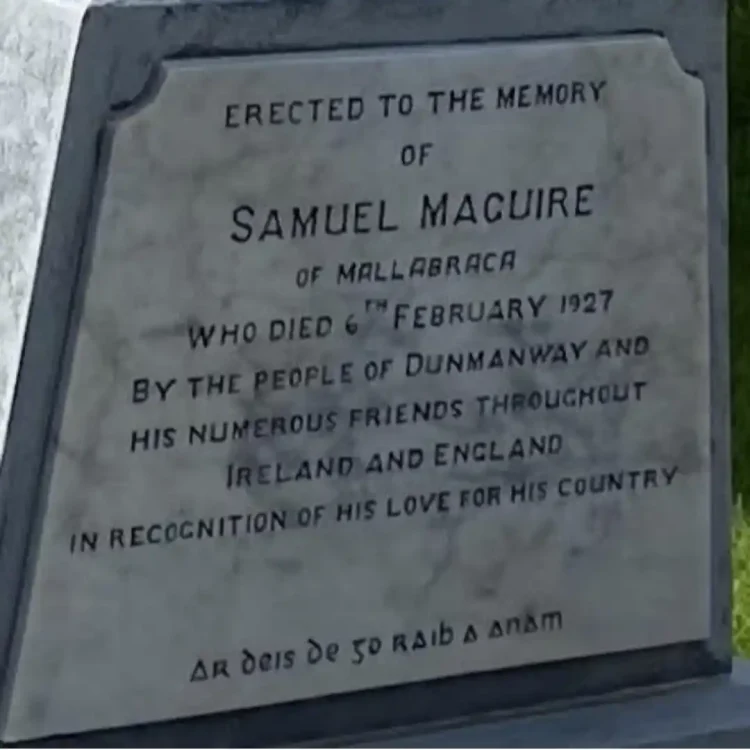 A close-up, tightly cropped photograph shows the rectangular white marble inscription plaque set into the tapered grey stone base of the Sam Maguire’s Grave. The stone has subtle grey veining running through it. The clean, serif capital letters are engraved directly into the stone and highlighted with dark paint for legibility. The bottom line of text is written in a traditional Irish uncial script. Transcribed Text ERECTED TO THE MEMORY OF SAMUEL MAGUIRE OF MALLABRACA WHO DIED 6TH FEBRUARY 1927 BY THE PEOPLE OF DUNMANWAY AND HIS NUMEROUS FRIENDS THROUGHOUT IRELAND AND ENGLAND IN RECOGNITION OF HIS LOVE FOR HIS COUNTRY. AR deis Dé go raibh a anam